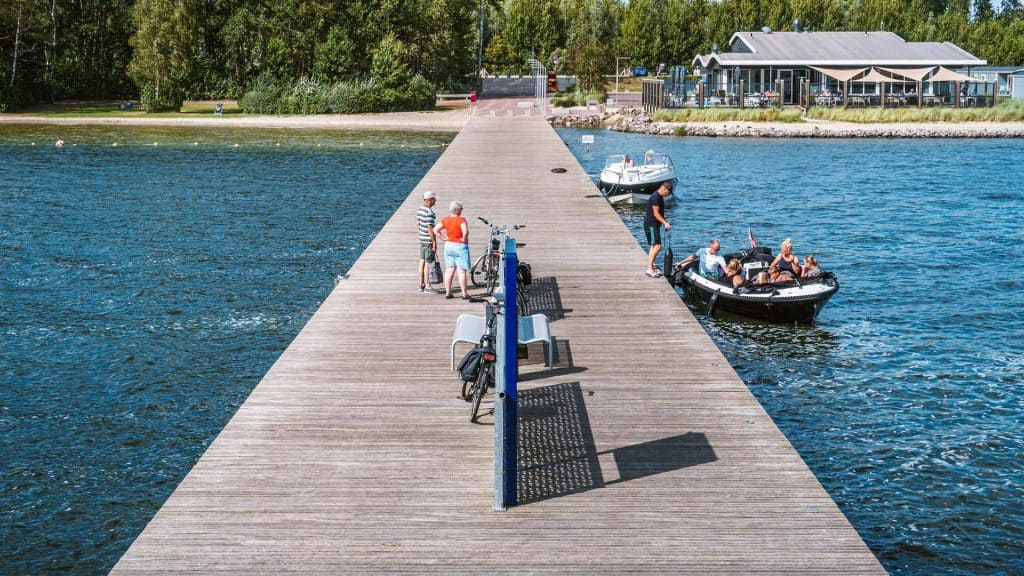 Foto van een steiger met aan beide kanten water. In het water liggen 2 bootjes. In 1 van de bootjes zitten mensen. Op de steiger staan 2 mensen en 2 fietsen.